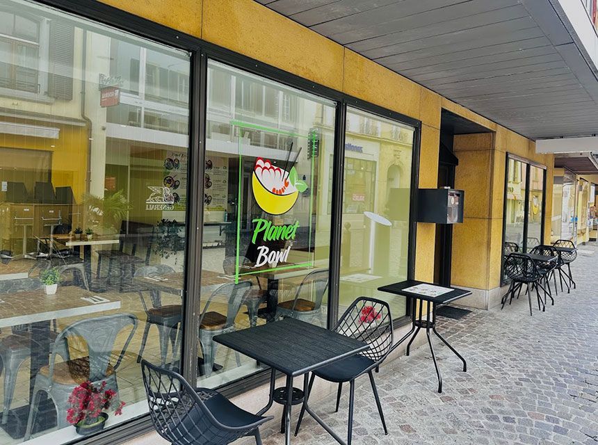 Un restaurant nommé « Planet Bowl » avec une terrasse et une façade vitrée. Des tables et des chaises noires sont installées sur le trottoir.