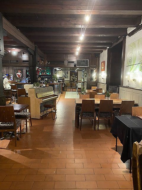 Restaurant intérieur avec piano, tables, chaises et sol en briques sous un plafond à poutres apparentes.