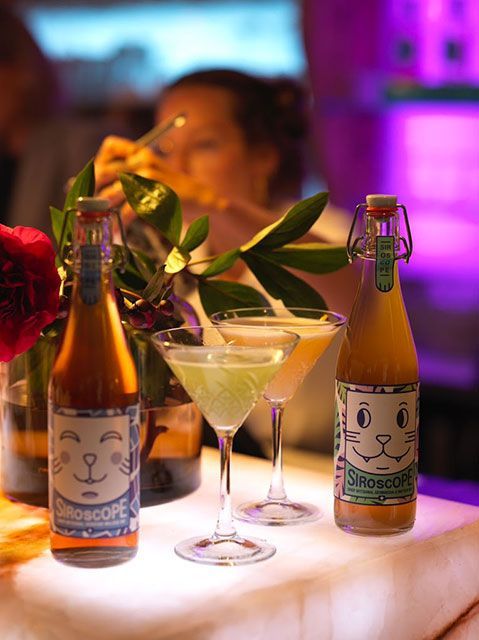 Deux bouteilles et des cocktails sont posés sur un bar. Une personne, à l'arrière-plan, mange avec des baguettes. Éclairage violet.