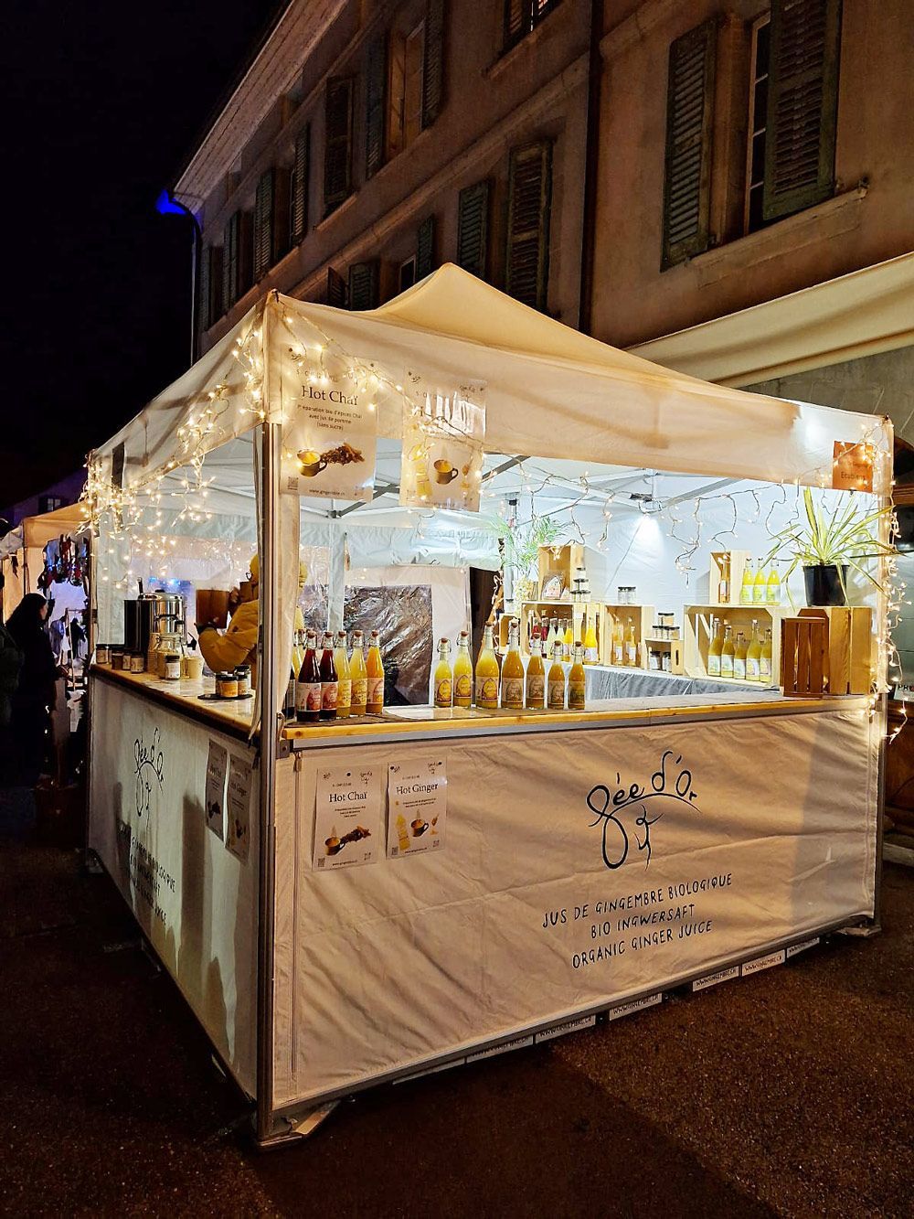 Étal de marché blanc la nuit, illuminé de guirlandes lumineuses, vendant de la nourriture et des boissons.