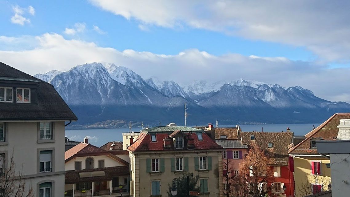 Bâtiments au premier plan, montagnes enneigées et lac sous un ciel bleu et nuageux.