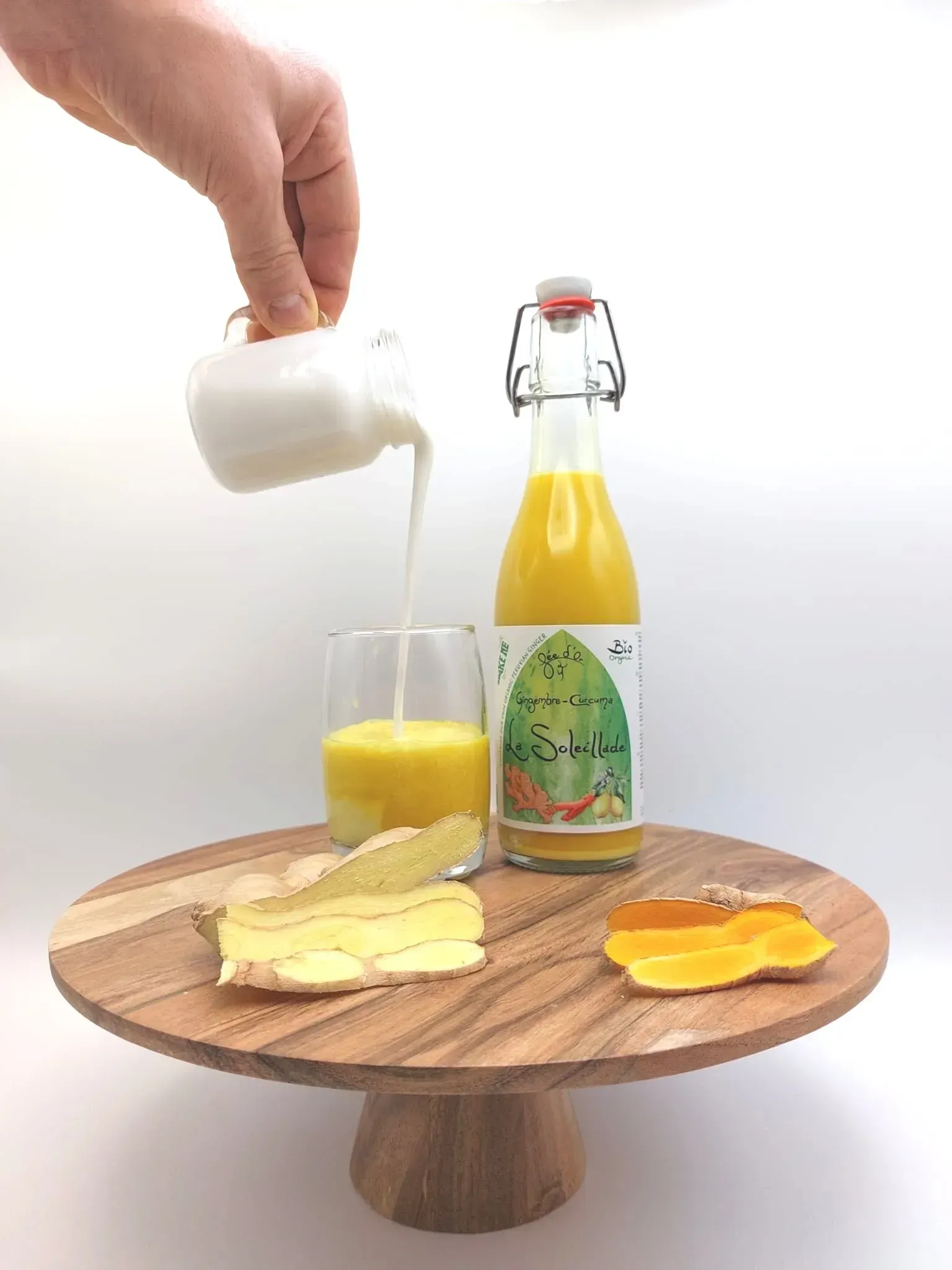 Verser de la crème à la main dans un verre de jus d'orange posé sur un présentoir à gâteaux en bois, à côté d'une bouteille de jus, de morceaux de fromage et de fruits.
