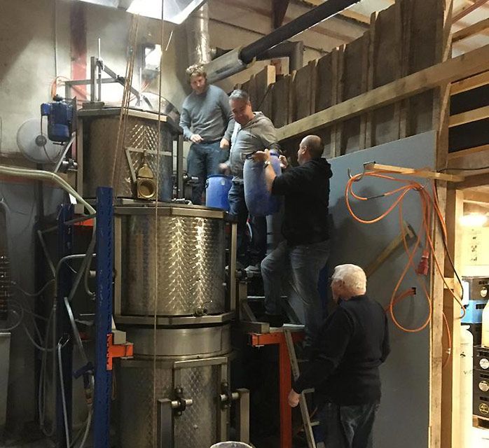 Des hommes travaillent dans une distillerie, transvasant du liquide de conteneurs dans des équipements industriels.