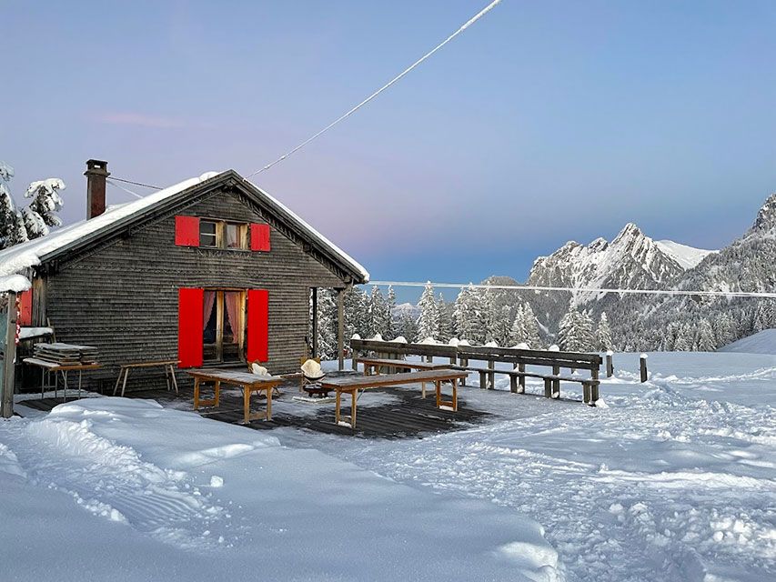 Cabane en bois enneigée avec volets et tables rouges, montagne en arrière-plan, paysage enneigé.