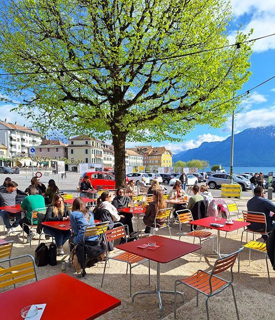 Café en terrasse avec tables rouges, clients, arbre et lac en arrière-plan.