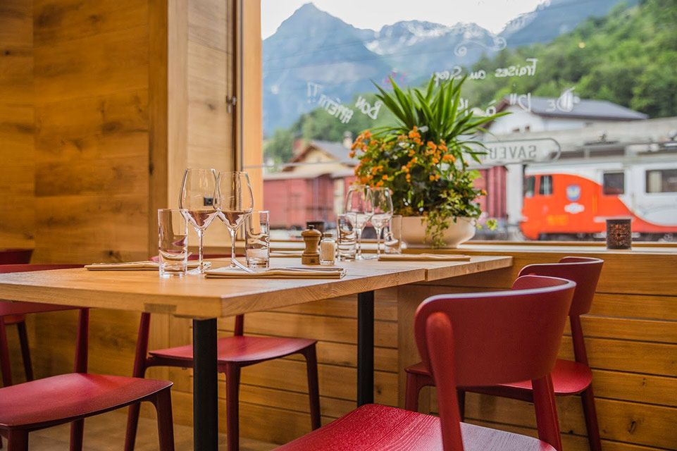 Table de restaurant avec chaises rouges, dressée pour le dîner, avec vue sur une montagne et un train.