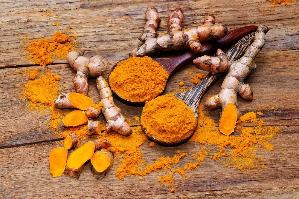 Racine de curcuma, tranchée et entière, avec deux cuillères en bois remplies de poudre de curcuma, sur une surface en bois.