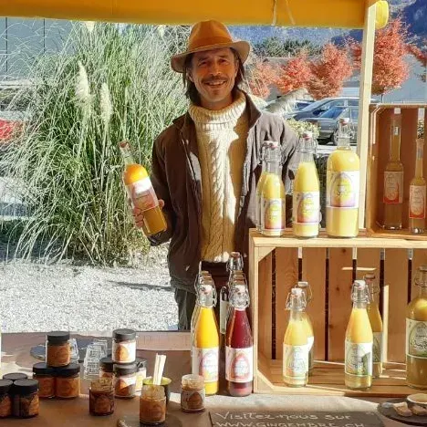 Un homme coiffé d'un chapeau vend du jus en bouteilles de verre sur un étal, par une journée ensoleillée.
