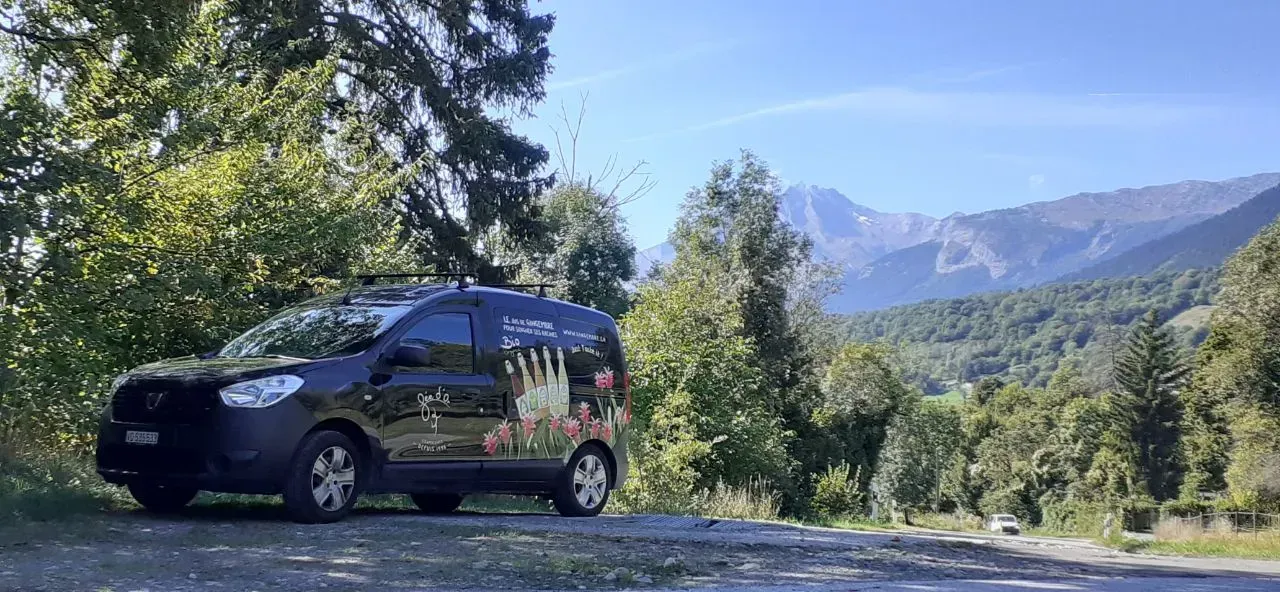 Une camionnette noire est garée sur une route avec un paysage montagneux en arrière-plan. Le véhicule est entouré d'un ciel lumineux et d'arbres.
