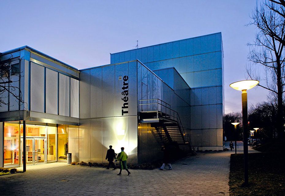 Façade d'un théâtre moderne au crépuscule, enseigne « Théâtre » visible. Deux personnes se dirigent vers l'entrée.