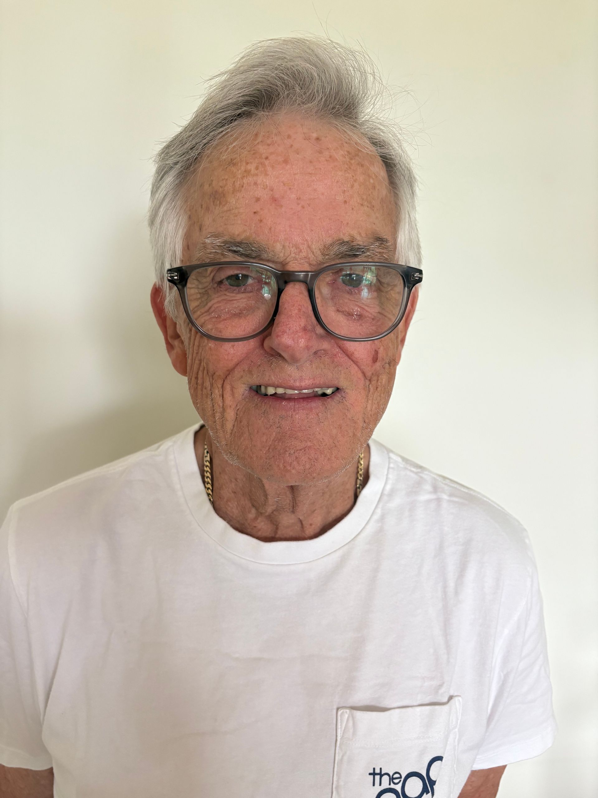 Smiling man wearing glasses and a white T-shirt against a plain beige wall