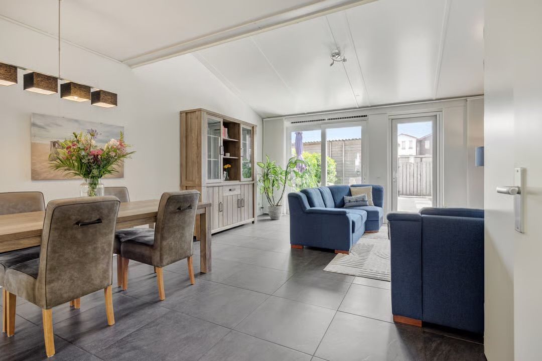 Na restyling, een moderne, minimalistische slaapkamer met een wit bed, grijze muren, een groot raam met uitzicht de tuin. De gekleurde kussen geven sfeer aan deze kamer.