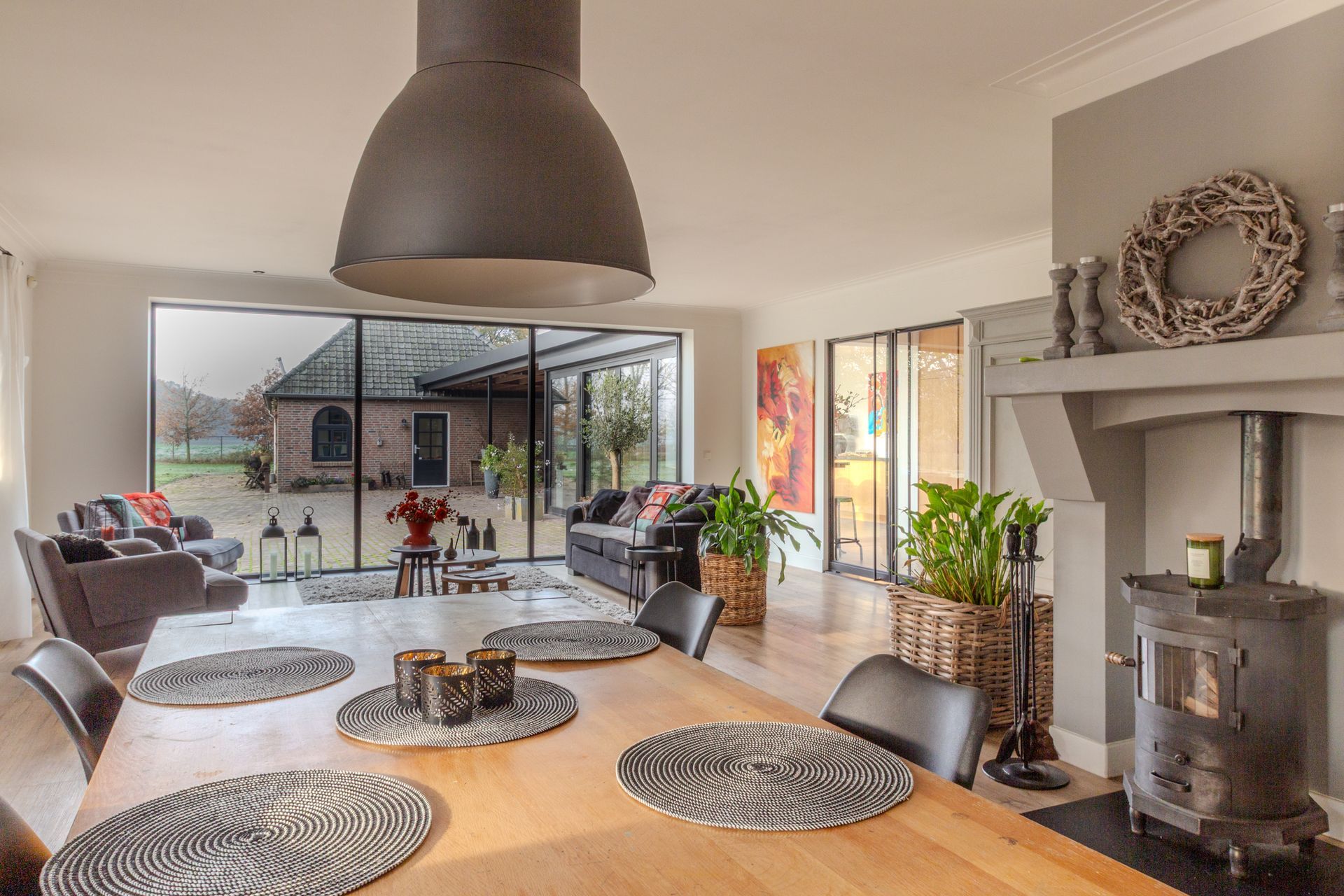 Een zonnige, open woonkamer met een grote houten tafel die dienst doet als extra eetplek, maar ook fungeert als heerlijke werkplek, autentieke gietijzeren openhaard en uitzicht op de tuin.