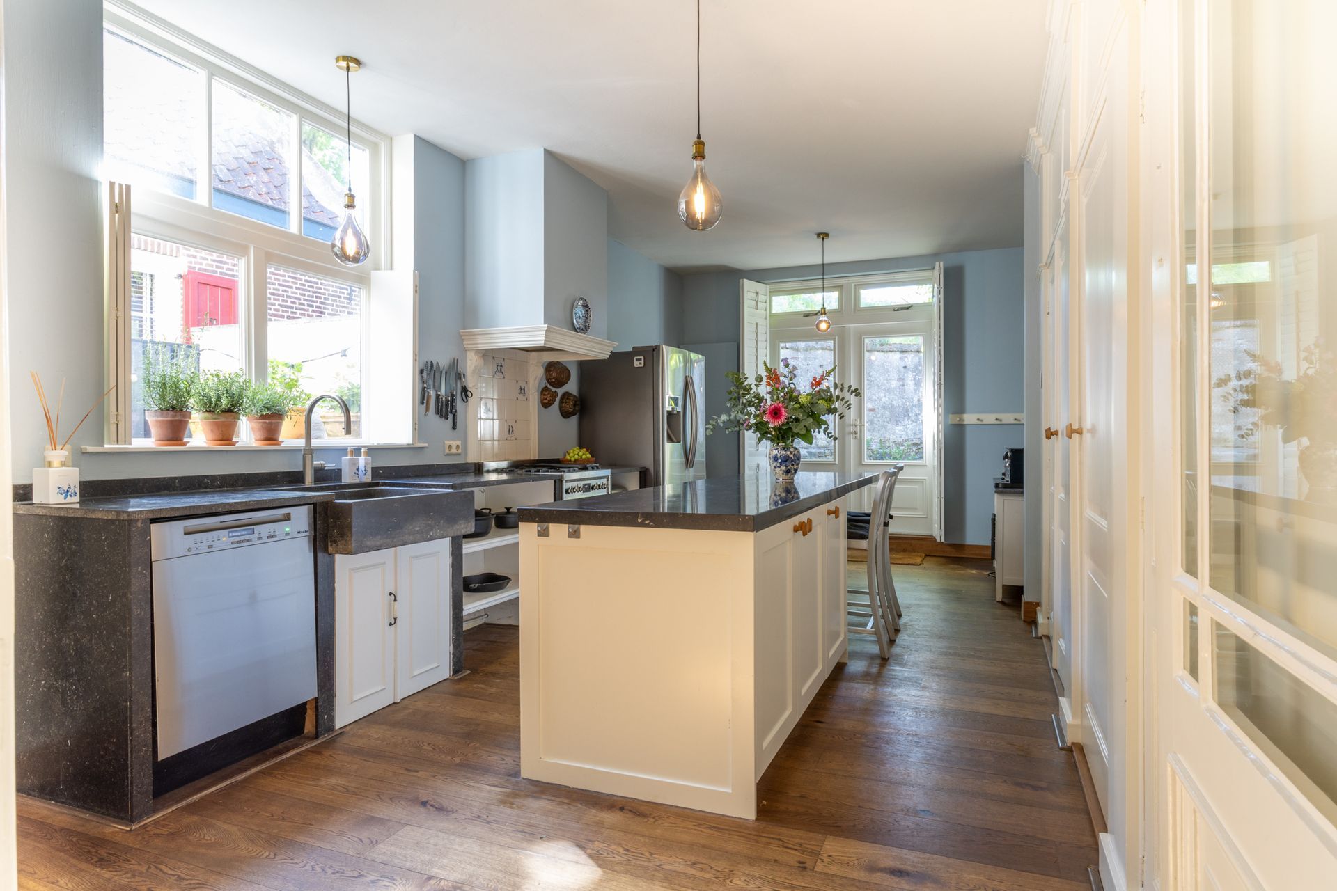 Na restyling, een moderne, minimalistische slaapkamer met een wit bed, grijze muren, een groot raam met uitzicht de tuin. De gekleurde kussen geven sfeer aan deze kamer.