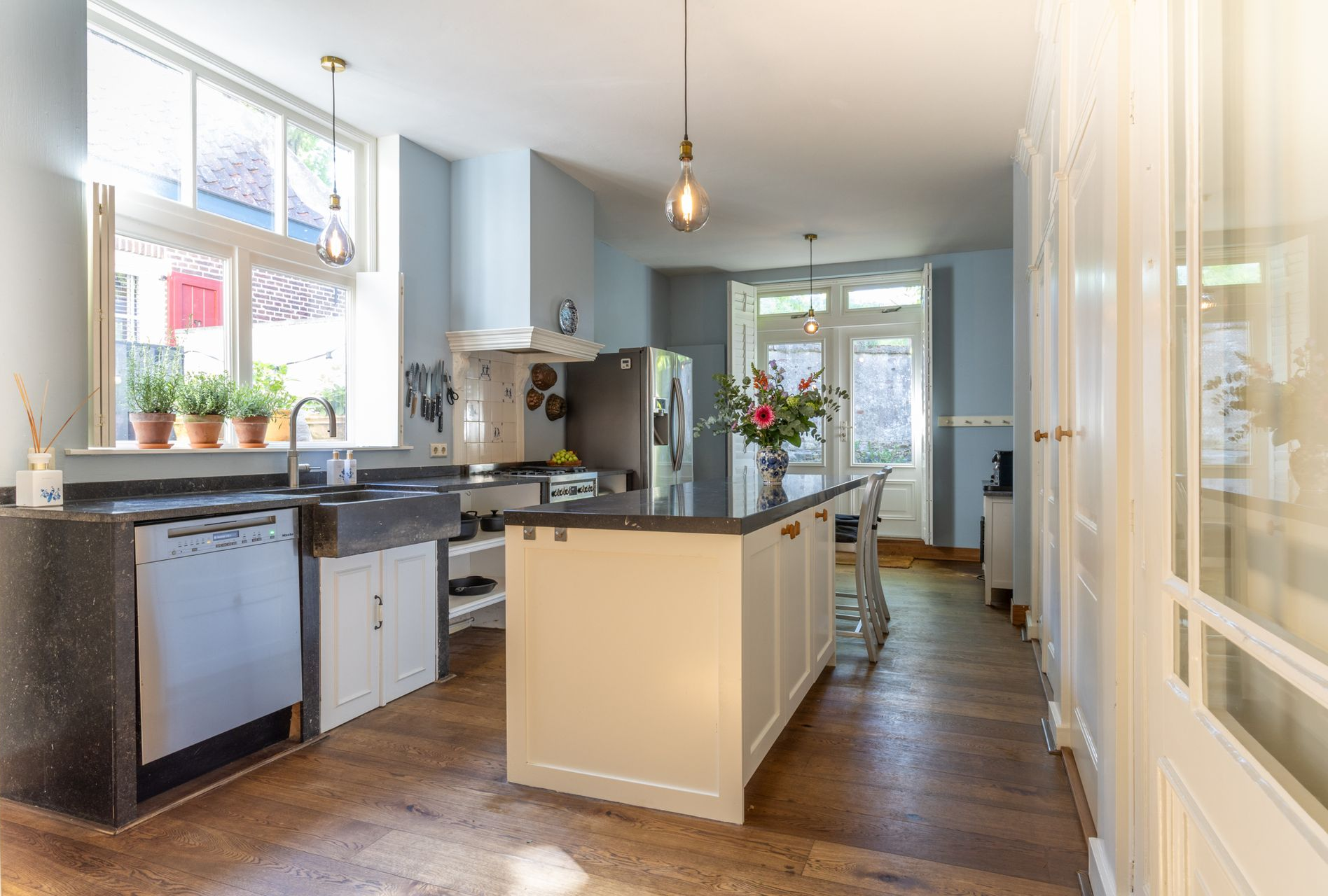 Na restyling, een moderne, minimalistische slaapkamer met een wit bed, grijze muren, een groot raam met uitzicht de tuin. De gekleurde kussen geven sfeer aan deze kamer.