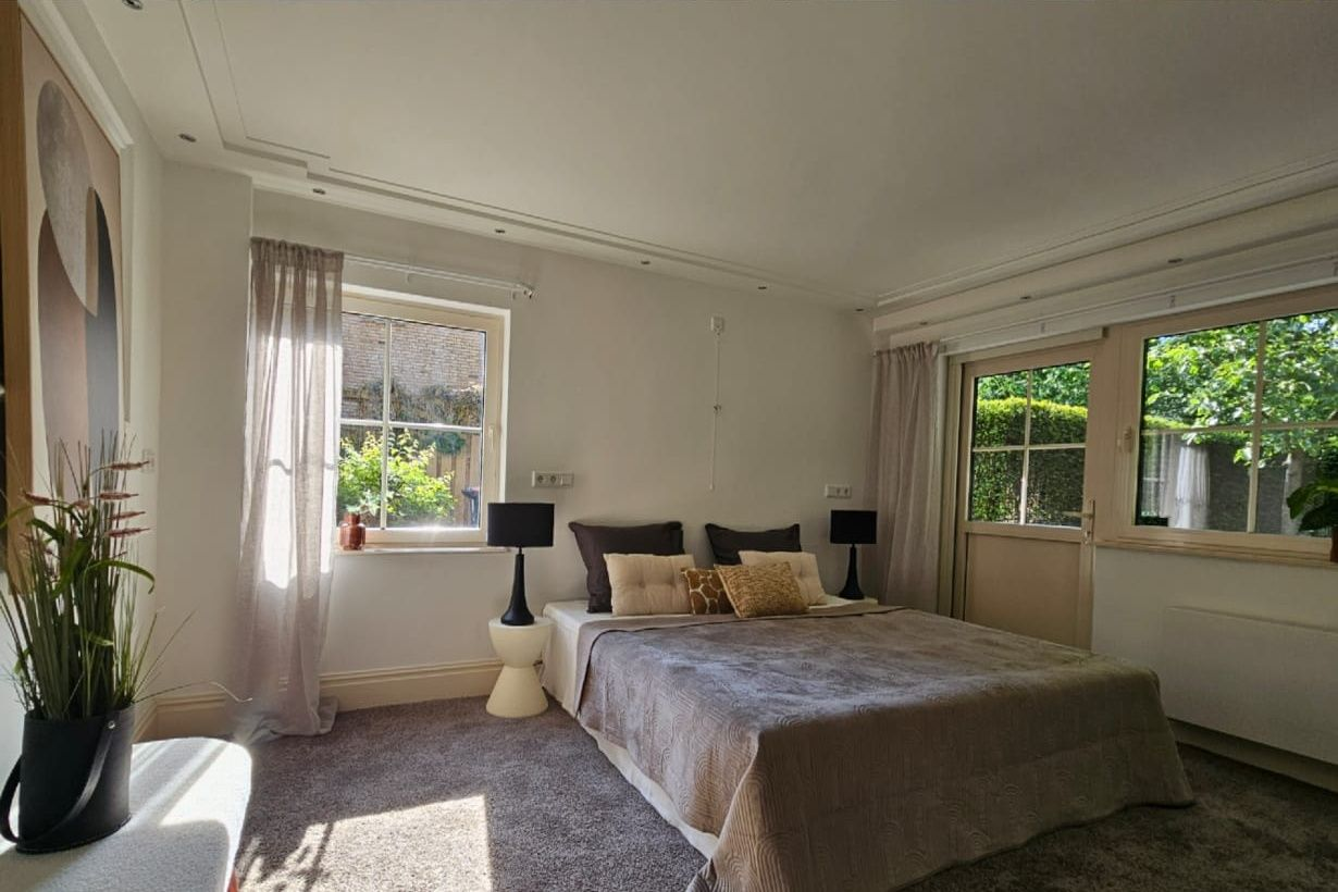 Na restyling, een moderne, minimalistische slaapkamer met een wit bed, grijze muren, een groot raam met uitzicht de tuin. De gekleurde kussen geven sfeer aan deze kamer.