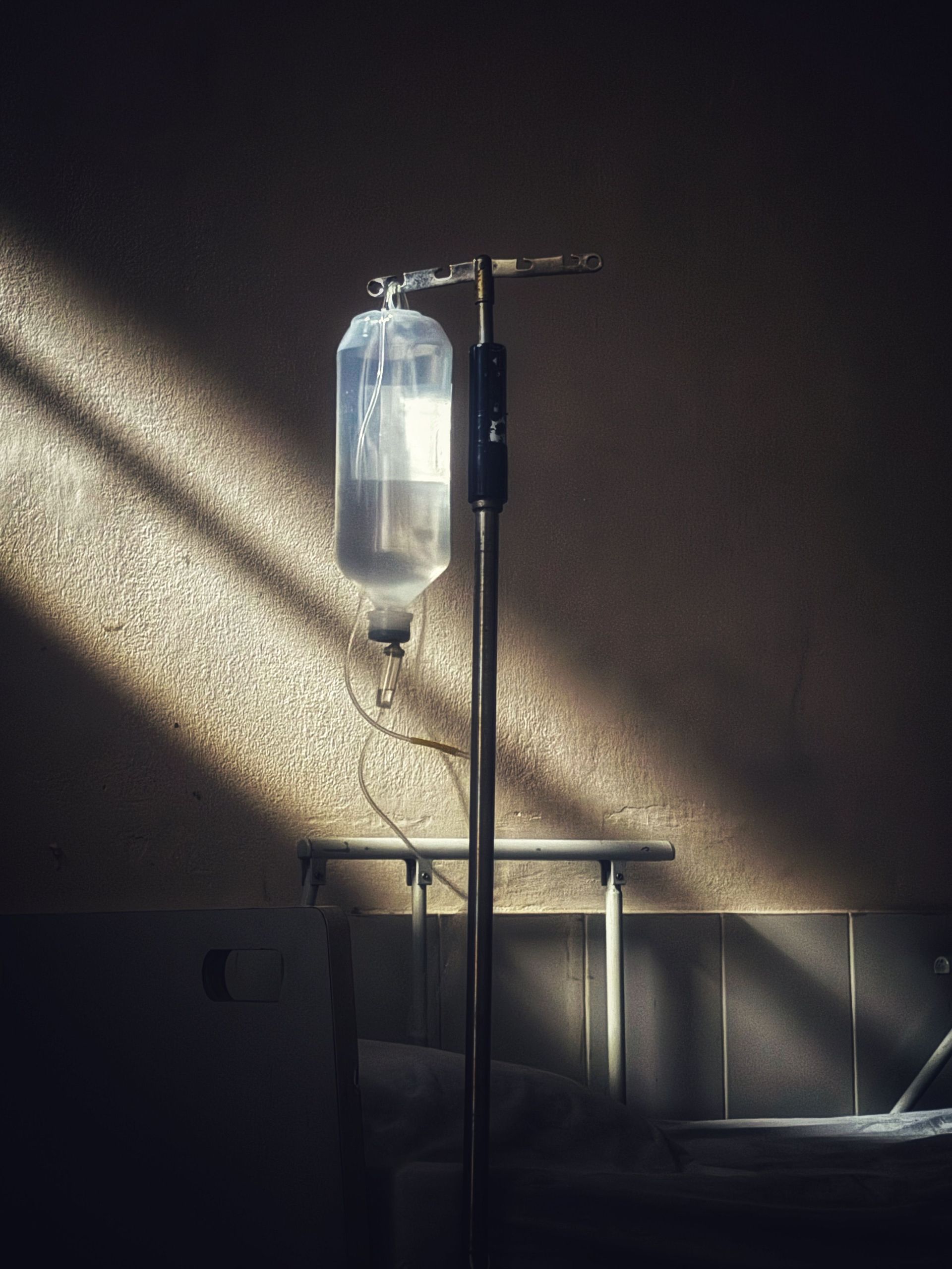 IV drip bag on a stand in a dim room, with sunlight streaming through blinds onto the wall