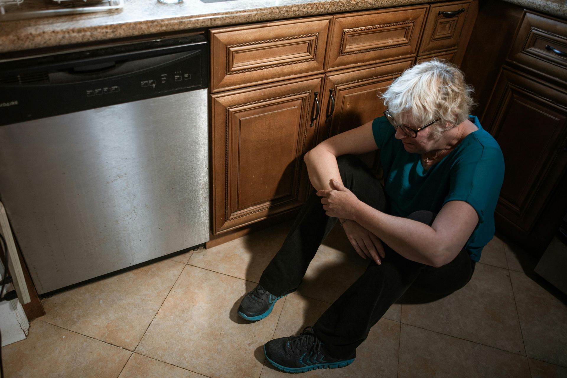 Woman sitting on kitchen floor next to dishwasher, looking down.