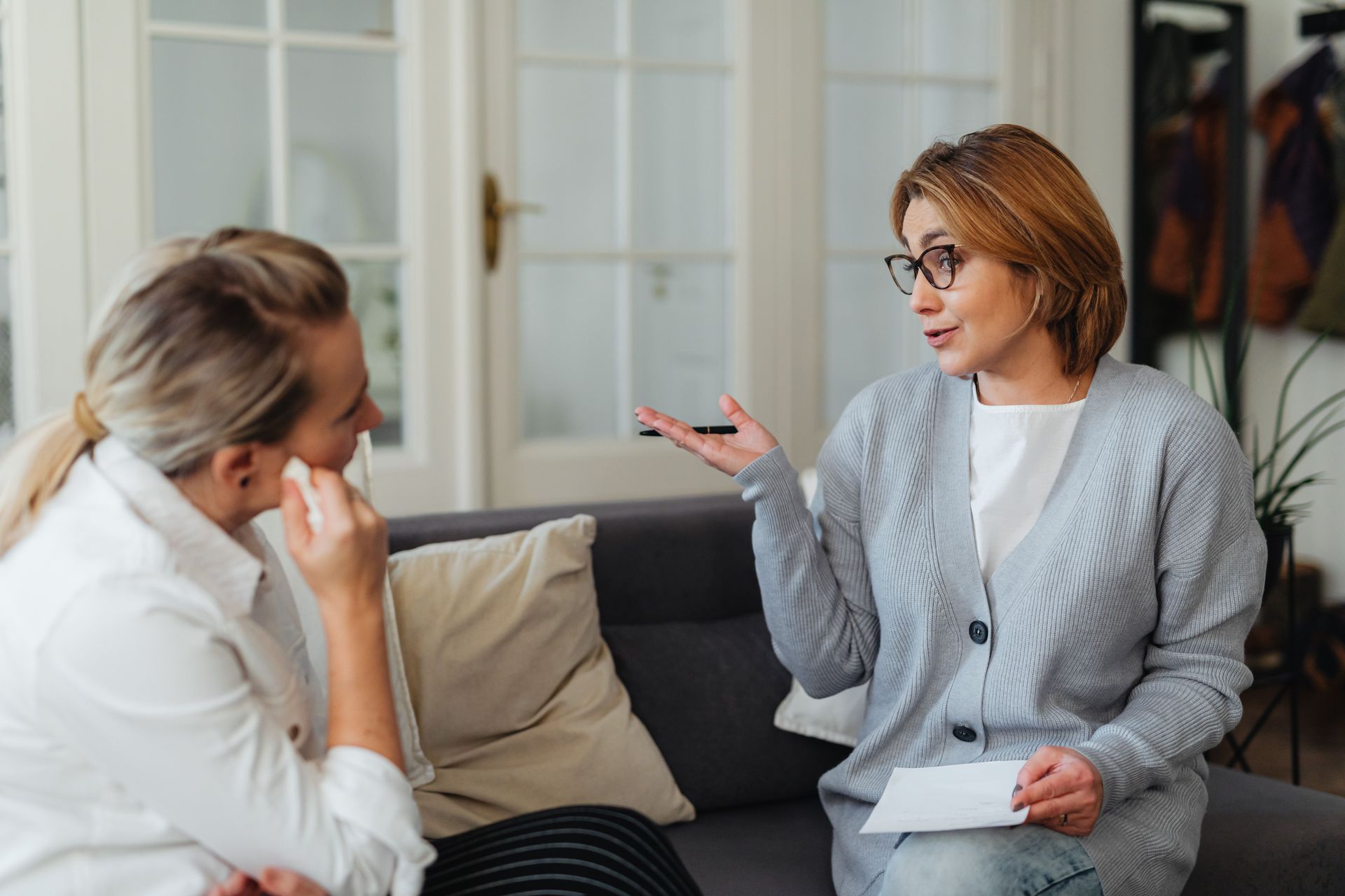 Two women talking on a couch; one gestures while holding a note in a bright living room.