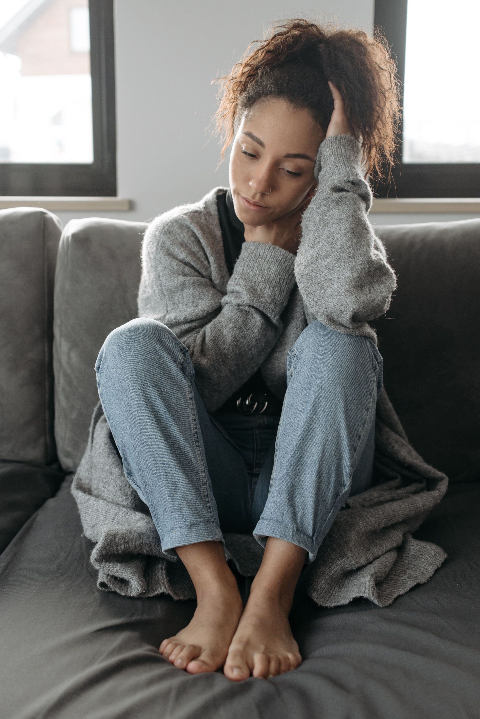 Woman sitting curled up on a gray couch, looking down with a pensive expression, in a softly lit room