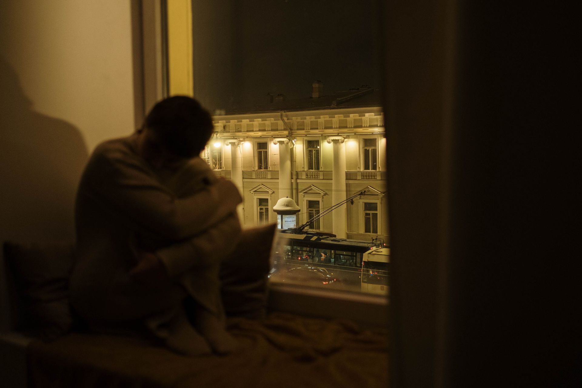 Person sitting by a window overlooking a lit building at night
