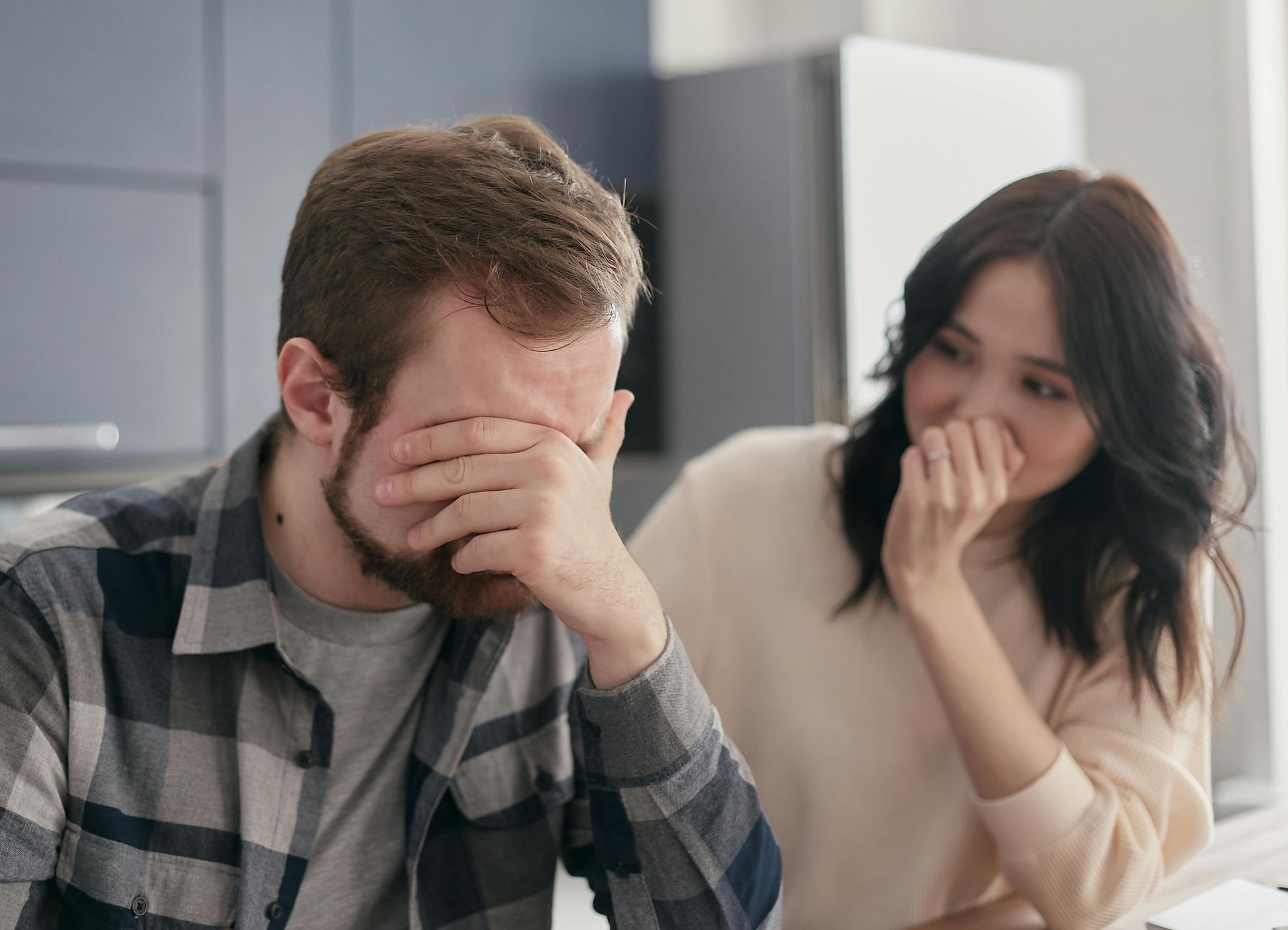 A person covering their face with their hands while sitting at a table near another person who is watching them.