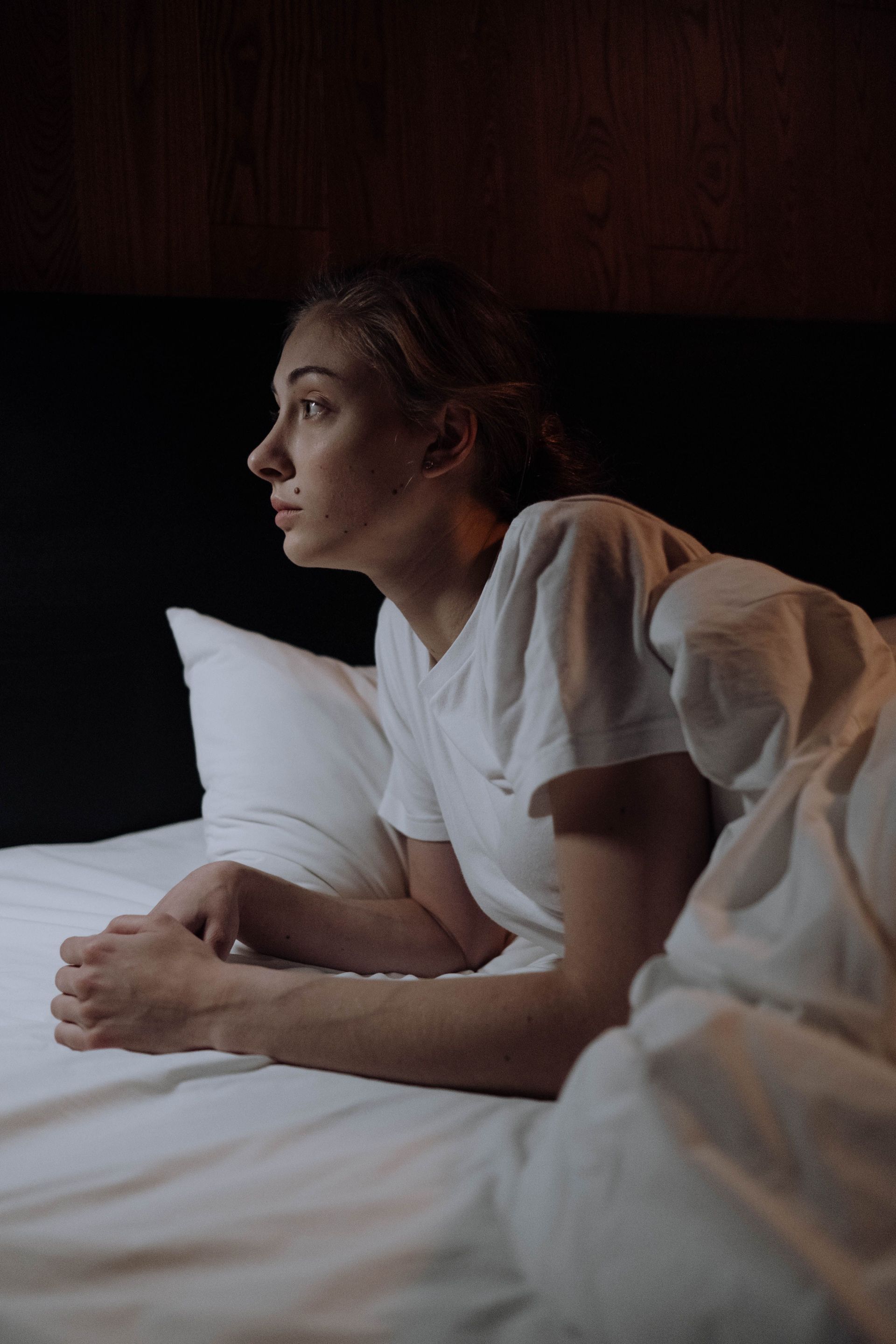 Person lying in bed, looking out thoughtfully in dim light, wrapped in white sheets.