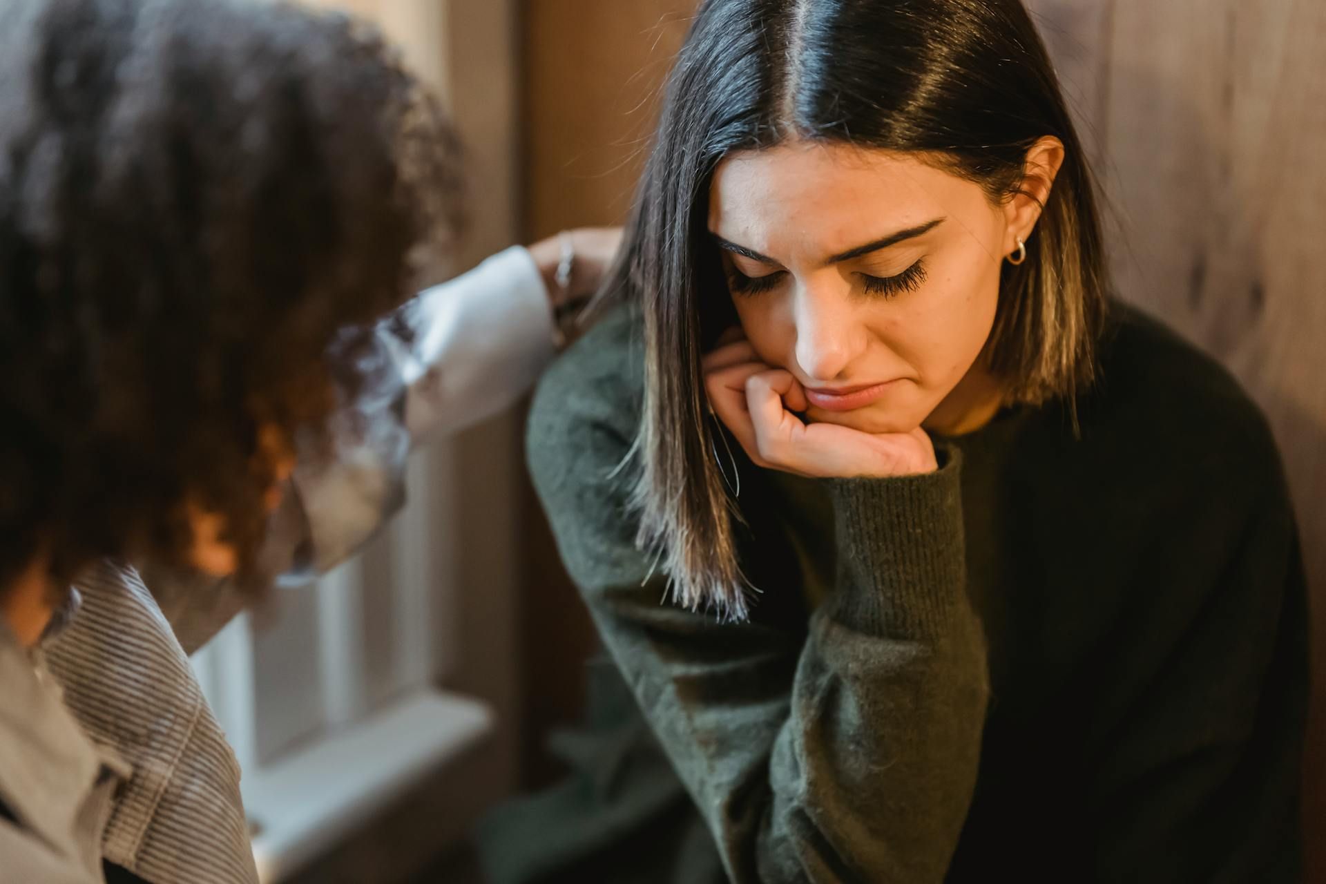 A person is comforting a woman with depression who is sitting next to a window.