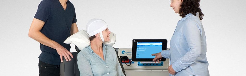a man and a woman are standing next to a woman sitting in front of tms therapy device and chair