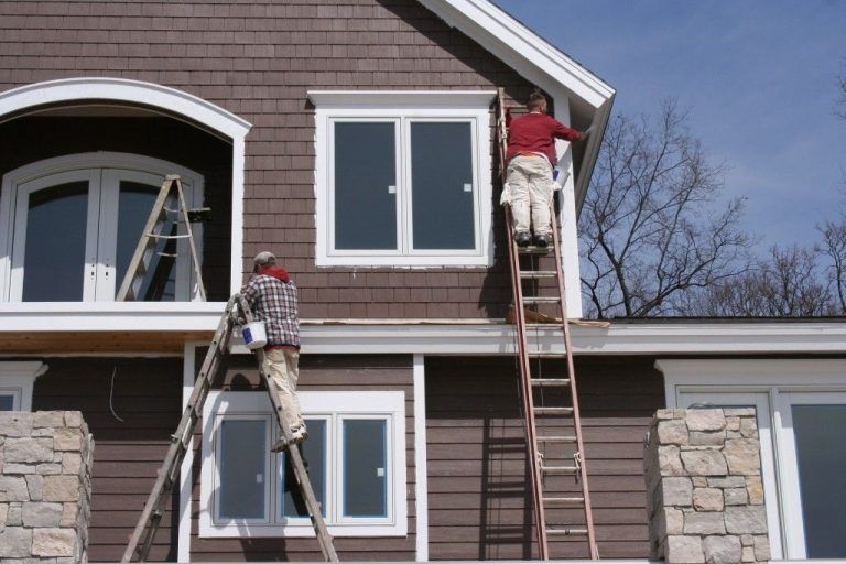Two workers painting a house exterior, one on a ladder and one at a window, with stone columns and brown siding.
