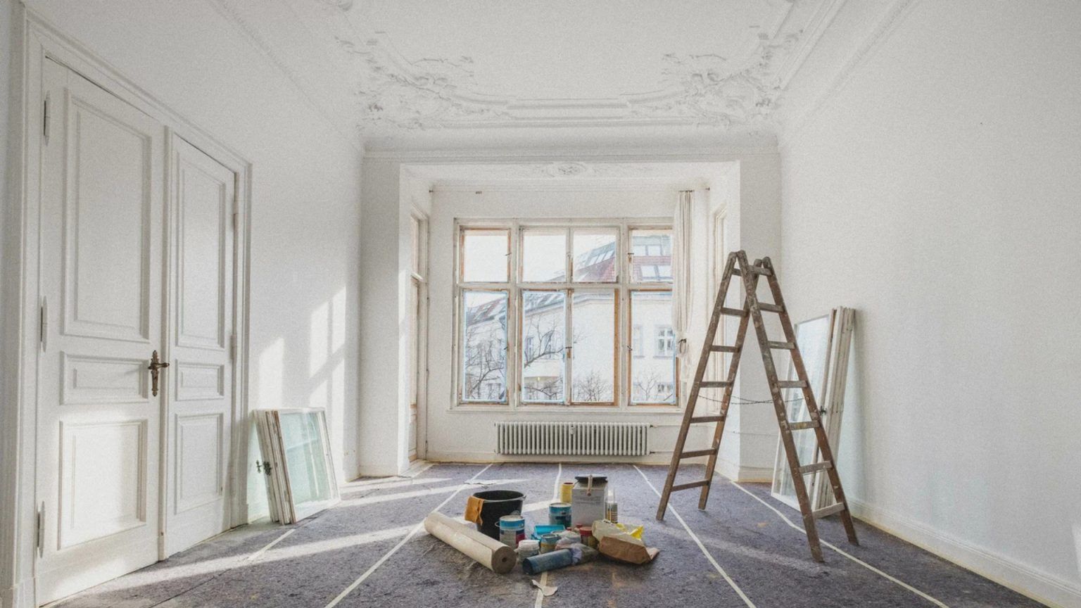 Bright empty room under renovation with ladder, paint supplies, and sunlight through a large window