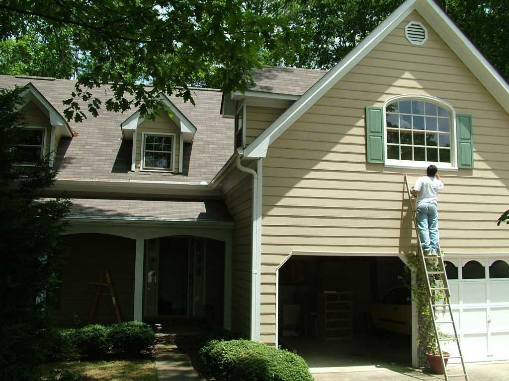 Person on a ladder painting the upper window trim of a beige suburban house