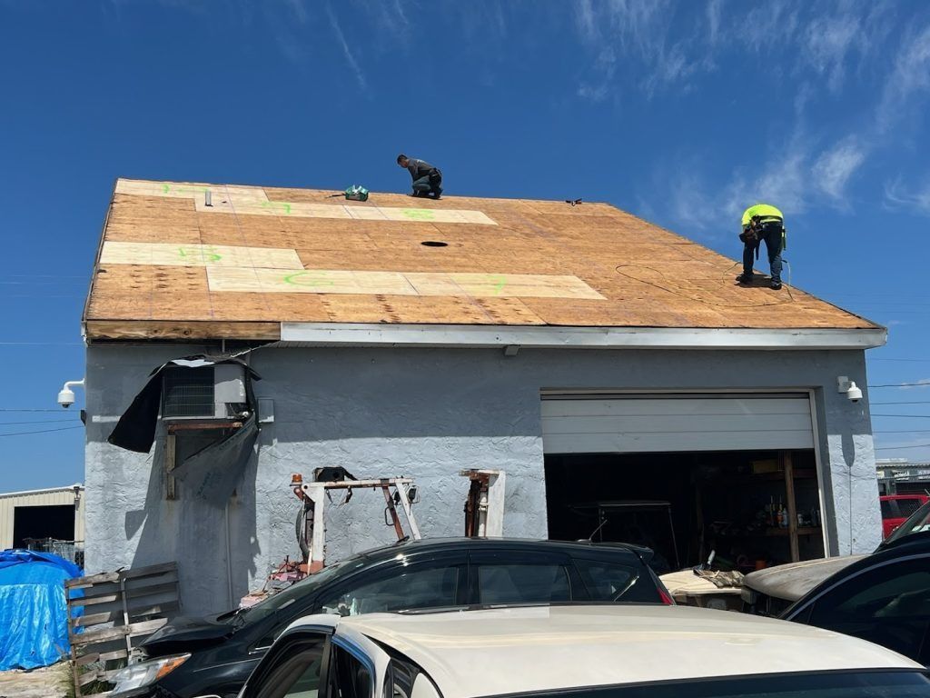 Workers roofing a gray garage, with plywood sheathing exposed and cars parked below.