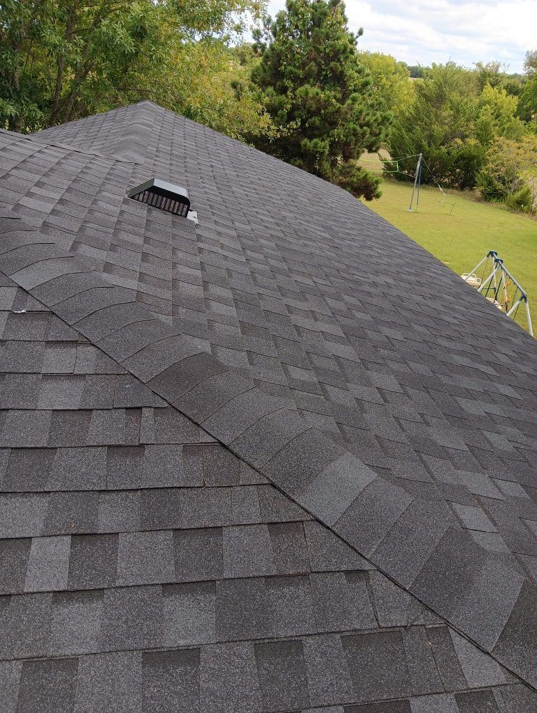 Roof with dark gray shingles and a vent, viewed from above with trees and lawn in the background