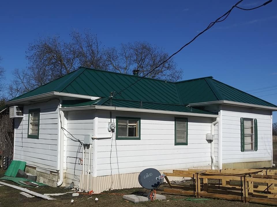White house with a green metal roof under a blue sky, with construction materials in the yard.
