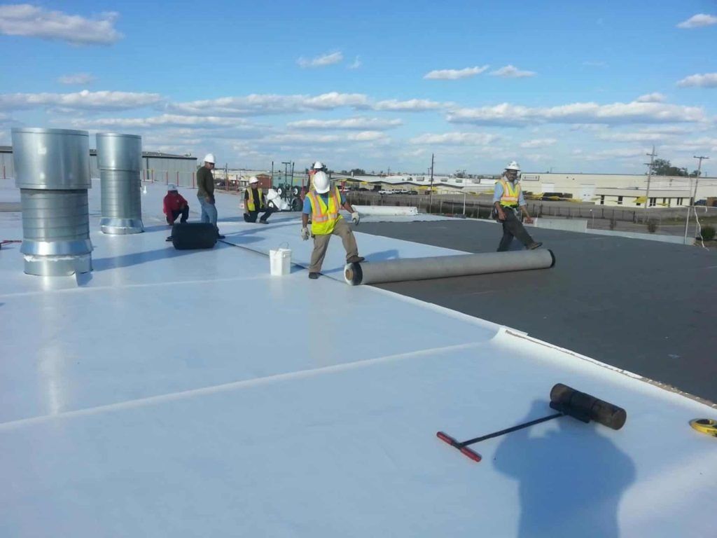 Workers installing roof membrane on a flat rooftop under a blue sky.