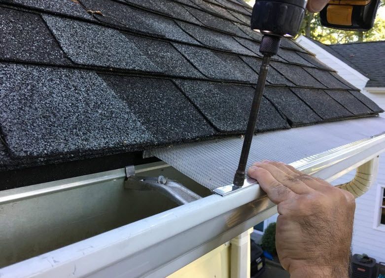Drill cleaning a clogged gutter beside a shingled roof, with a hand steadying the gutter edge.
