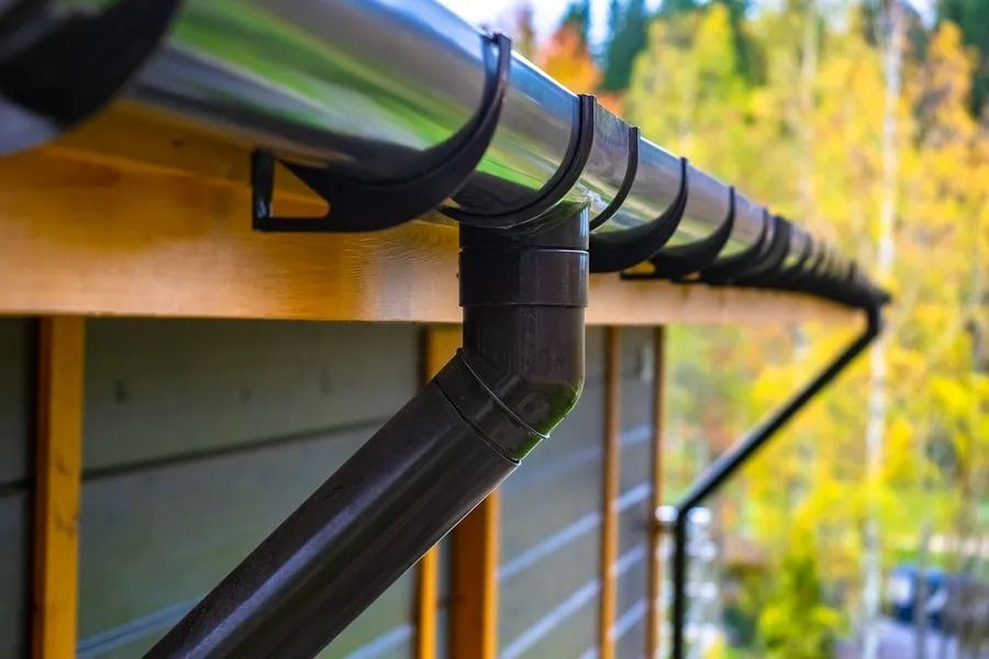 Black gutter and downspout on a house roof with autumn trees in the background