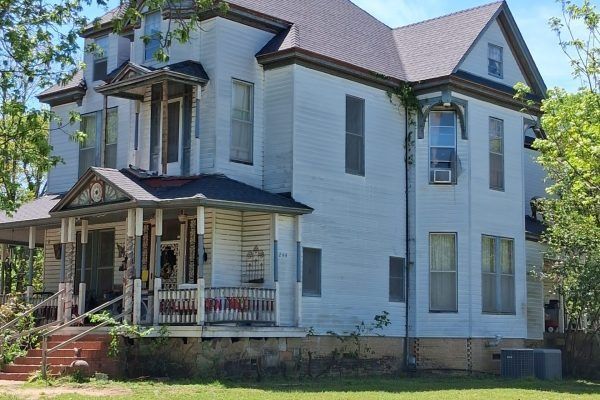 White two-story Victorian house with a porch, gabled roof, and front yard surrounded by trees