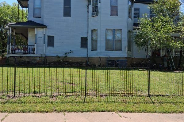 White house with a front porch behind a black metal fence and grassy yard along a sidewalk.