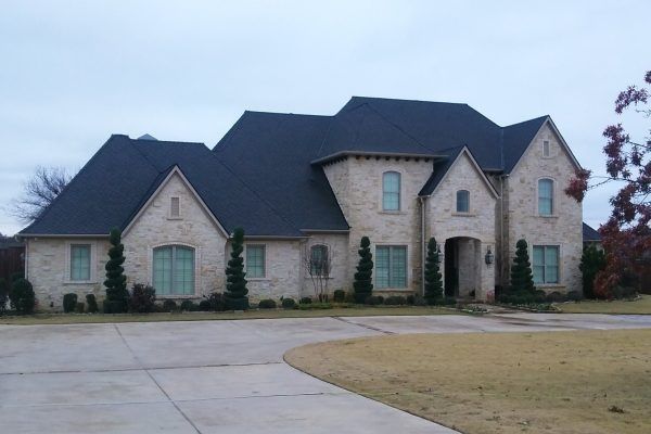 Large stone suburban house with dark roof, arched windows, and a wide driveway under a cloudy sky