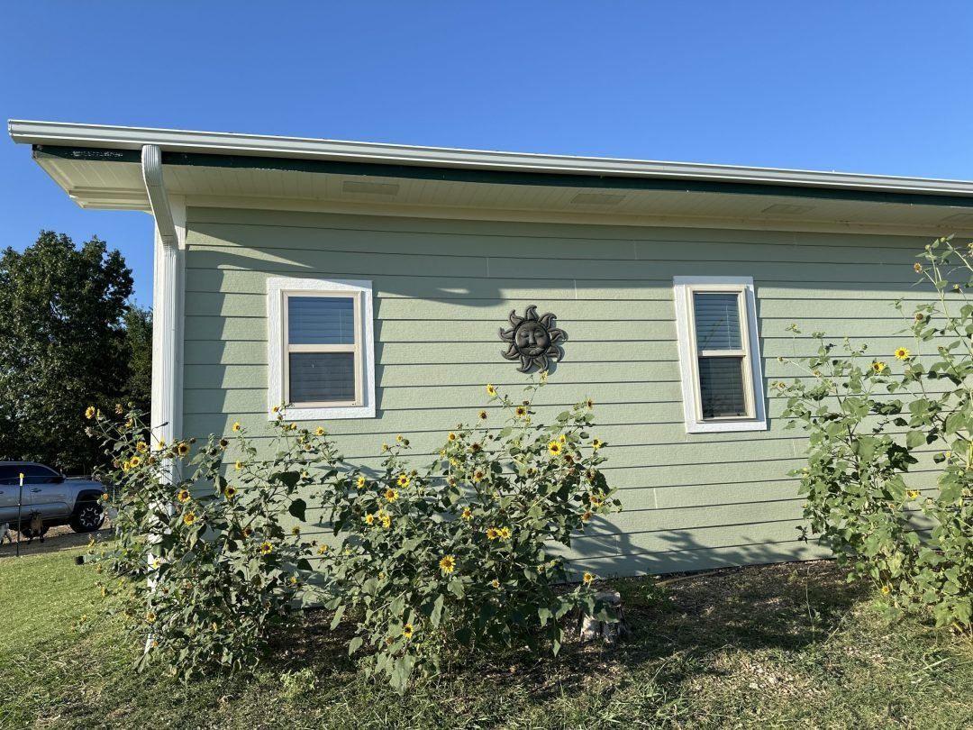 Light green house exterior with two white-trimmed windows and shrubs in front under a blue sky