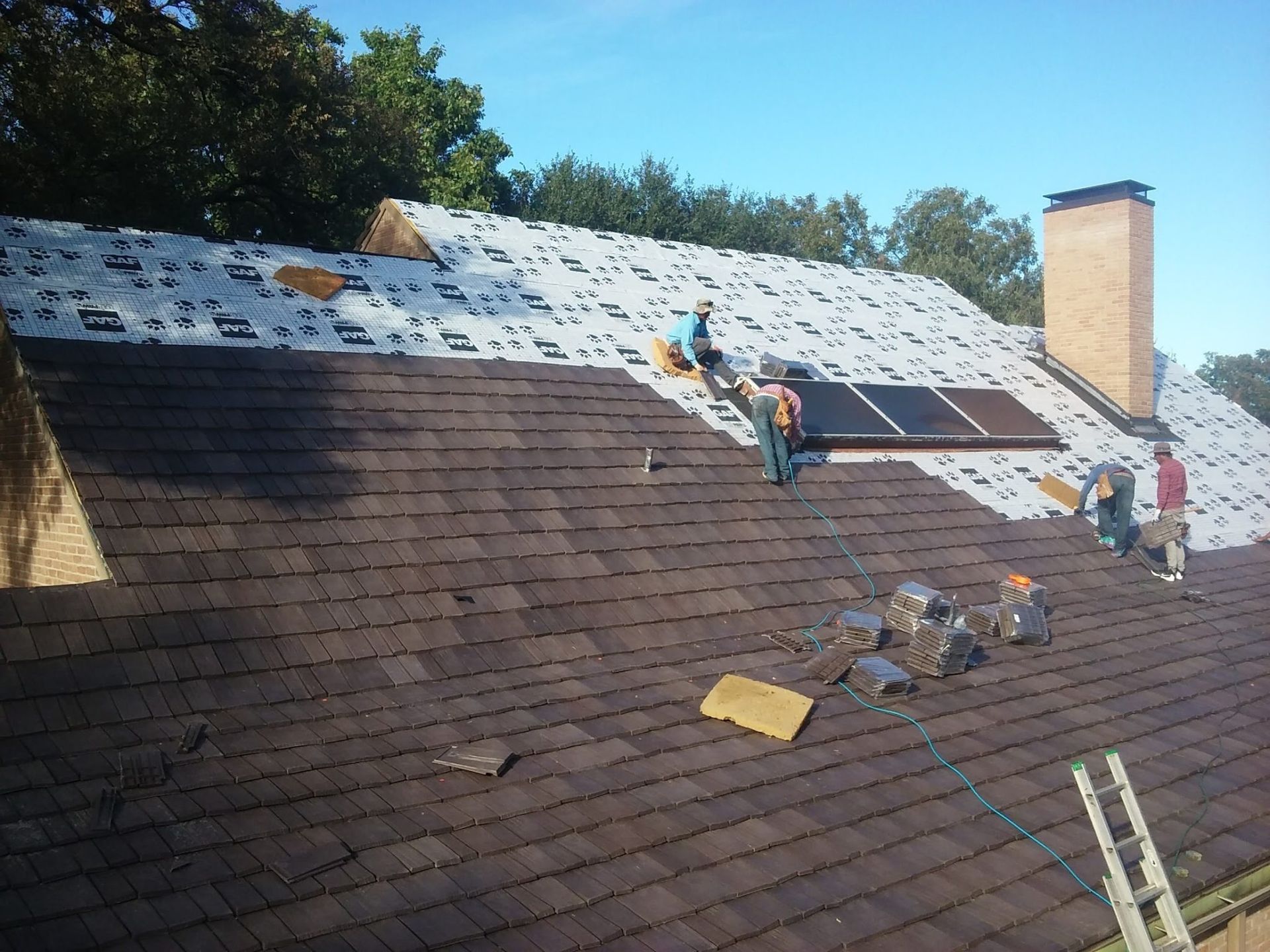 Roofers repairing a shingled roof, with exposed underlayment and a chimney under a blue sky.