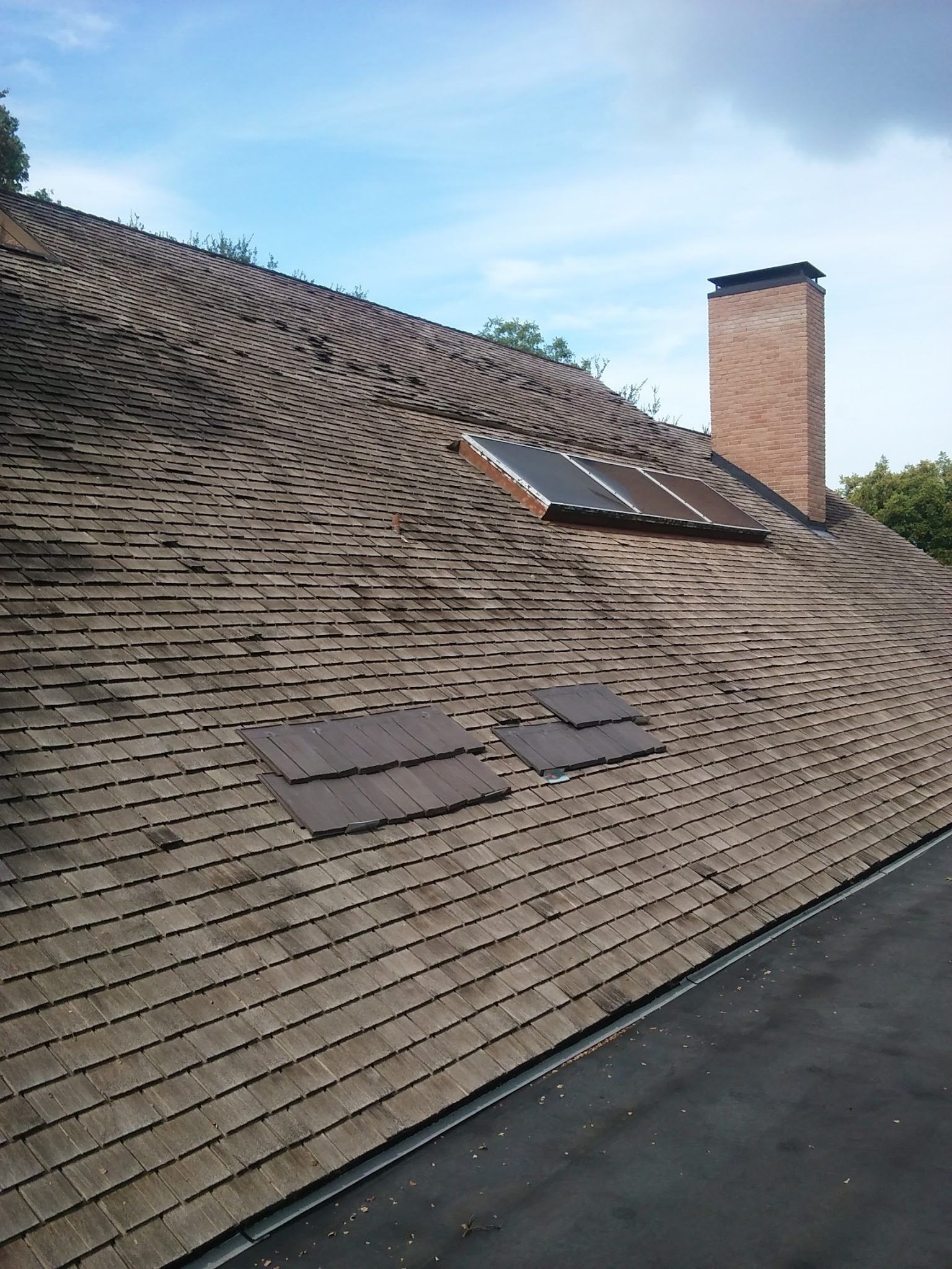 Shingled roof with several damaged patches and a chimney under a clear sky