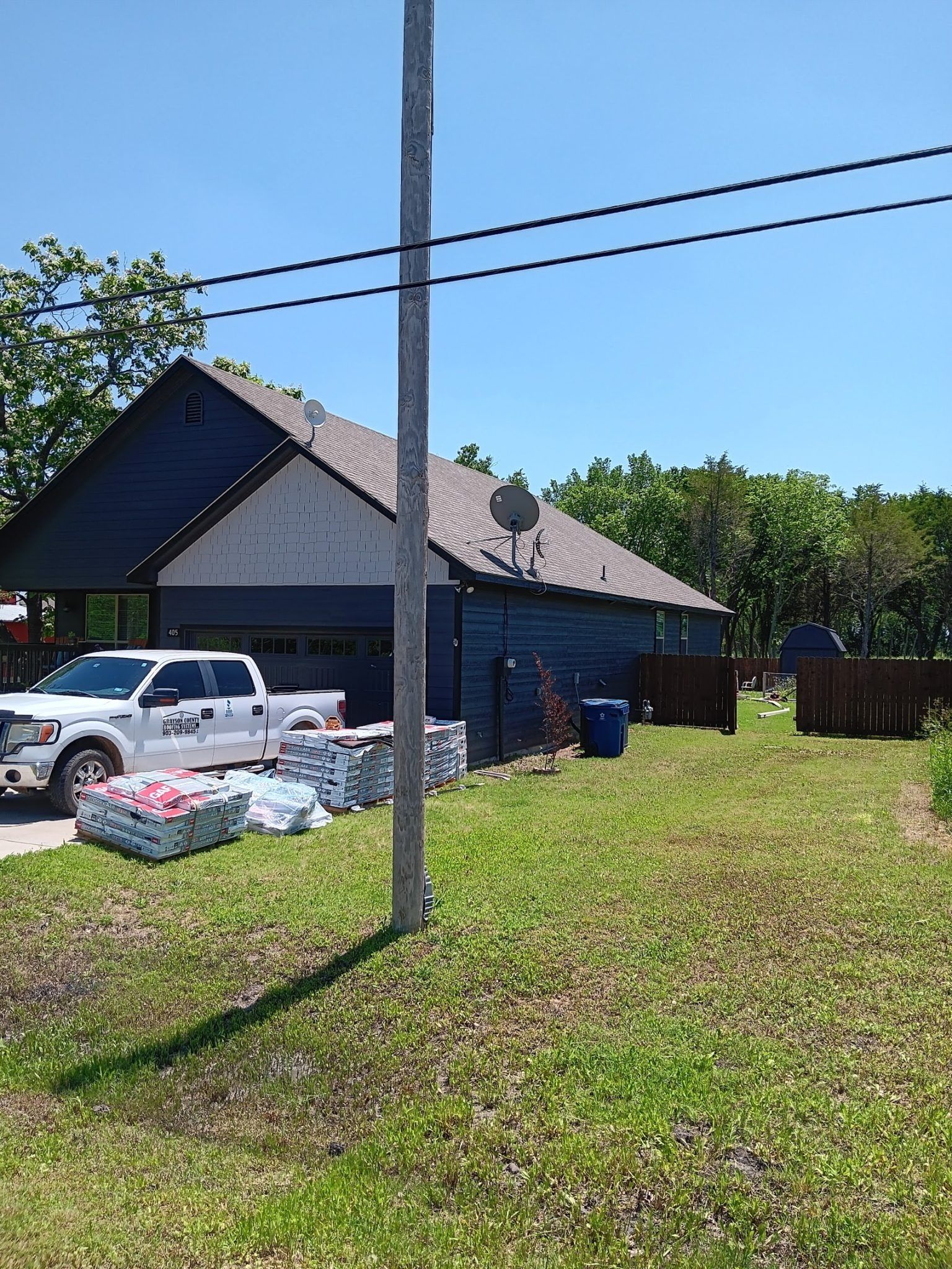 Single-story dark house with parked vehicles and yard, seen from across a grassy lot under a clear blue sky