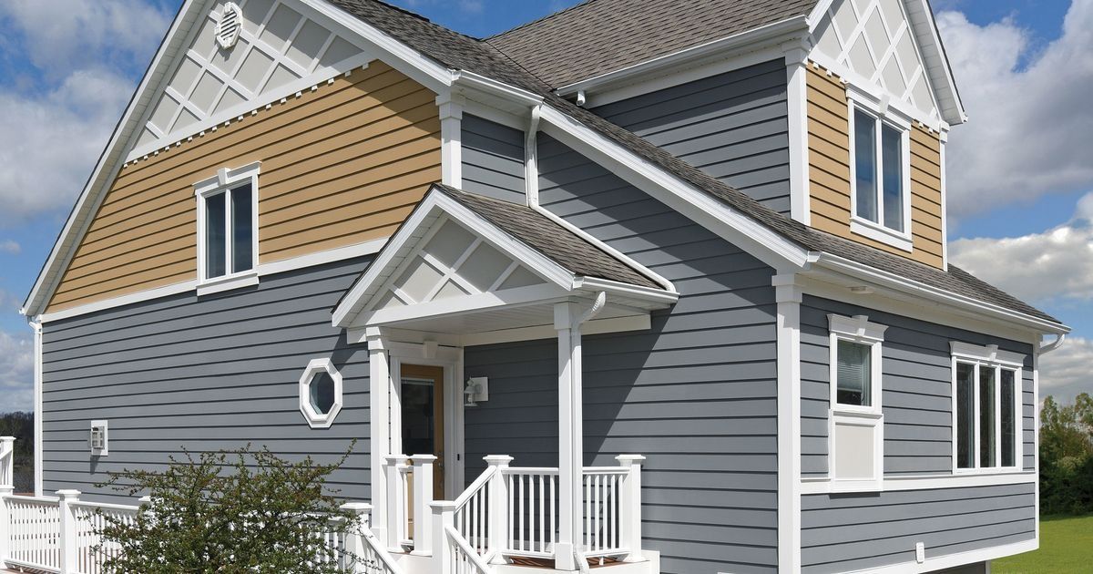 Two-story gray and tan house with white trim, front porch, and dormer windows under a cloudy sky