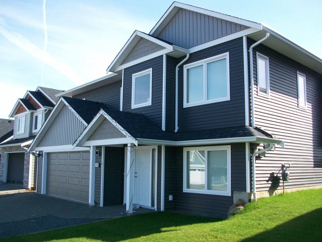 Row of dark gray two-story suburban townhouses with garages and green lawn under a bright sky