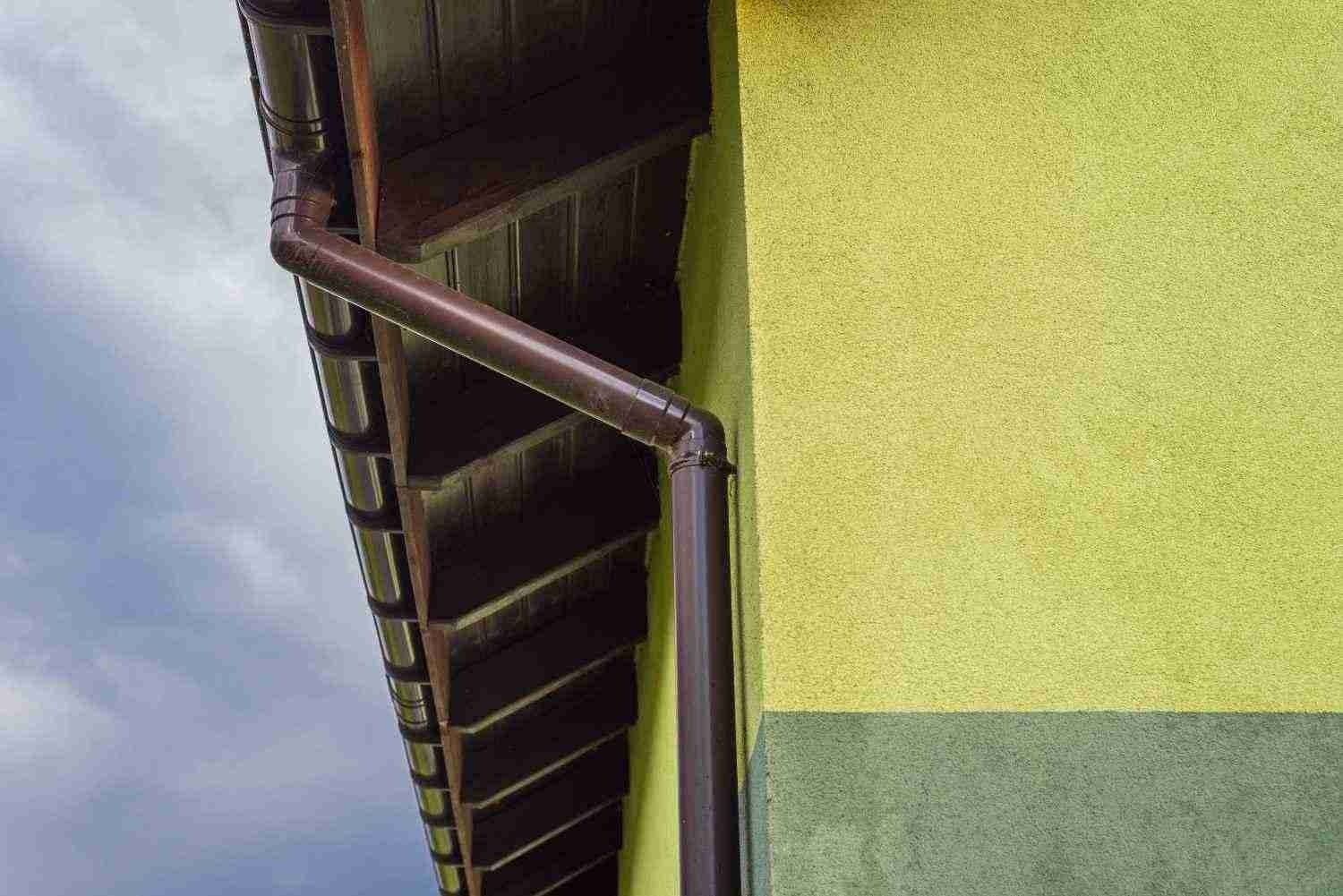 Rain gutter on a house roof beside a yellow wall and cloudy sky