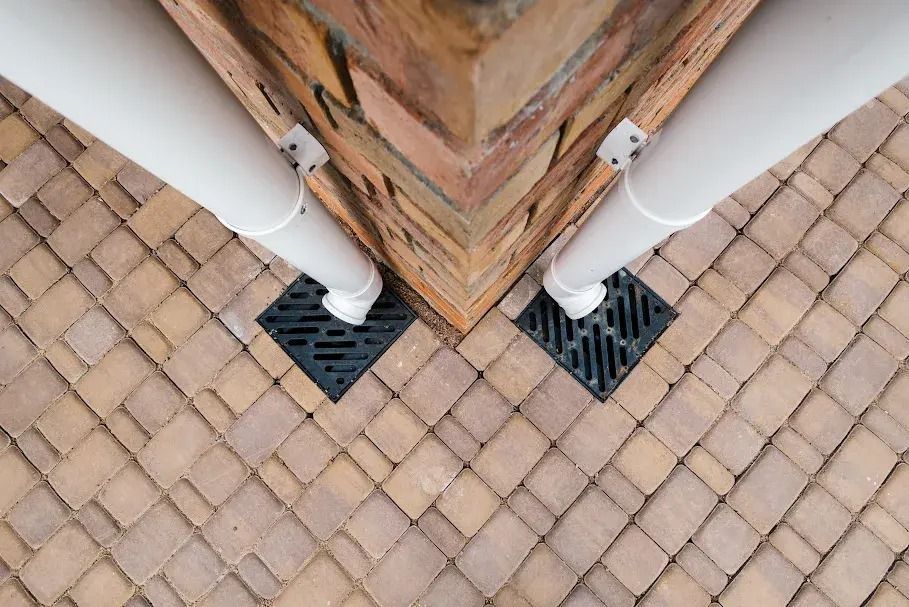 Two white downspouts meeting at a brick corner above black drain grates on a tiled patio