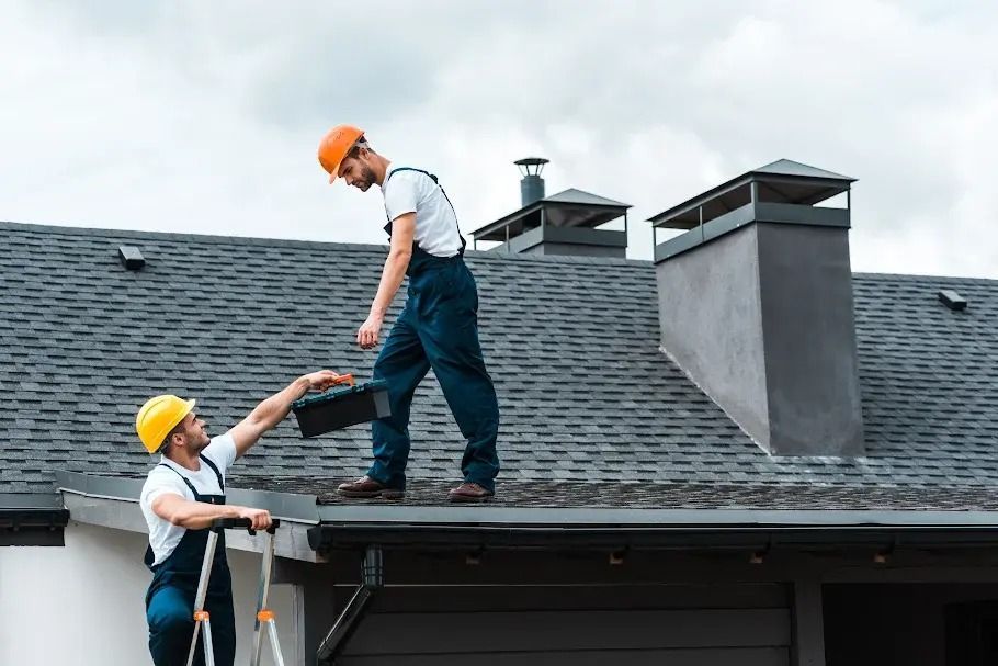 Two roofers on a gray shingle roof, one handing tools to the other near a chimney.